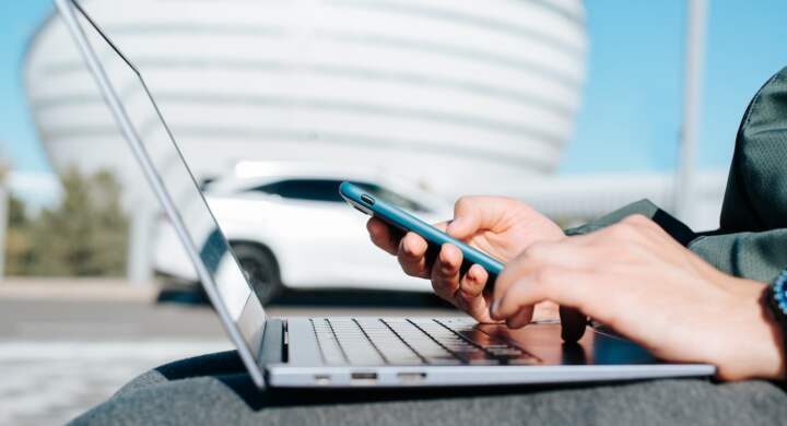 Close-up of man sitting in business district and working remotely on laptop, typing on keyboard and browsing internet on smartphone. Selective focus on hand with mobile phone, side view.