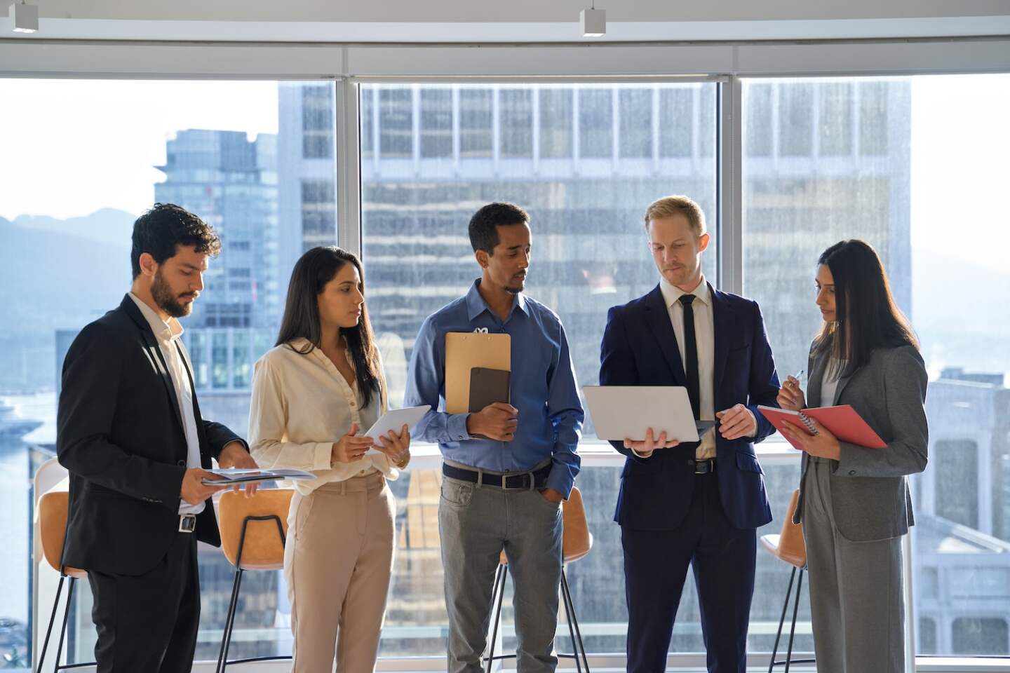 Young happy diverse professional team business people standing in modern office and talking. International workers group having teamwork discussion planning strategy with devices at work meeting.