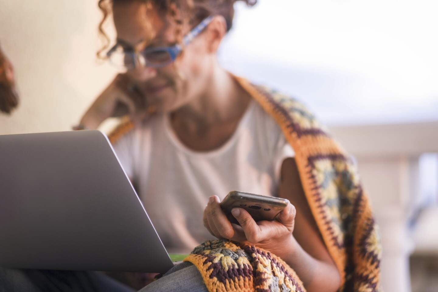 Young woman holding mobile phone while working on laptop. Woman working on laptop sitting at house terrace. Woman pleased while holding mobile phone and reeiving good news using laptop
