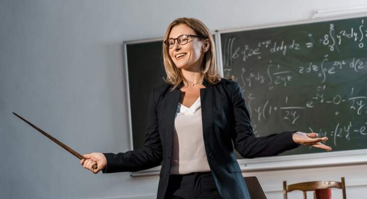 smiling female teacher with wooden pointer explaining mathematical equations in classroom