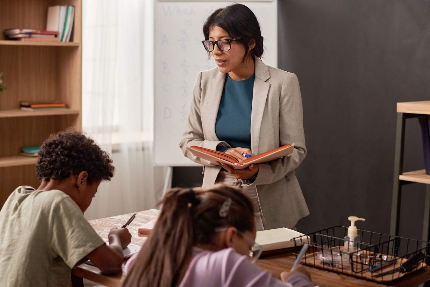Serious Hispanic teacher in glasses standing with book and holding dictation while children writing it at class