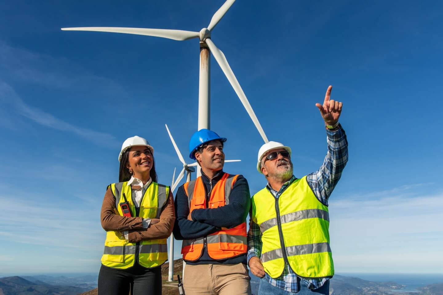 Three multiethnic engineers wearing safety vests and helmets are standing in a wind farm, with one of them pointing towards a wind turbine, discussing renewable energy generation