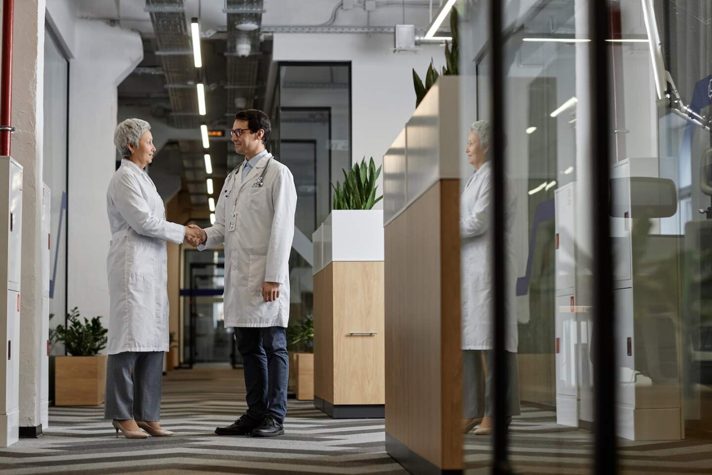 Two mature intercultural doctors in lab coats shaking hands and looking at one another while standing in hospital corridor and making agreement