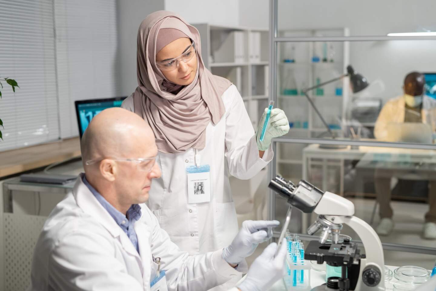 Young serious Muslim female clinician in hijab with flask communicating with mature male colleague pointing at tablet screen