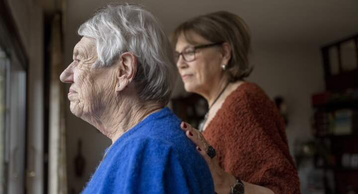 Two elderly friends standing looking out of the window consoling one another