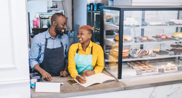 Charming African American couple in yellow and gray shirts and blue and black aprons writing orders at counter and looking with pleasure
