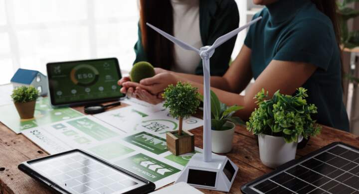 Two businesswomen holding a green globe and discussing an esg project with miniature wind turbine and solar panel on a wooden table