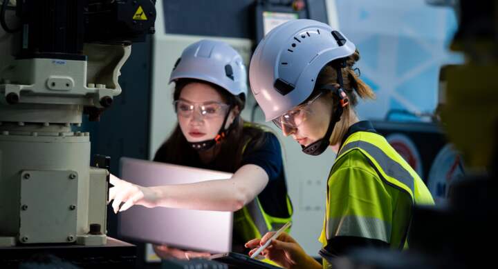 Two women wearing safety gear and helmets are working on a computer. One of them is writing on a piece of paper